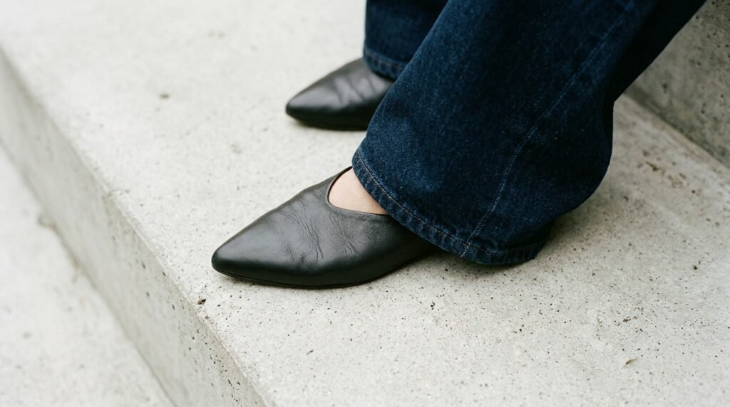 Pointed-toe leather flats resting on a concrete step next to the hem of wide-leg jeans.